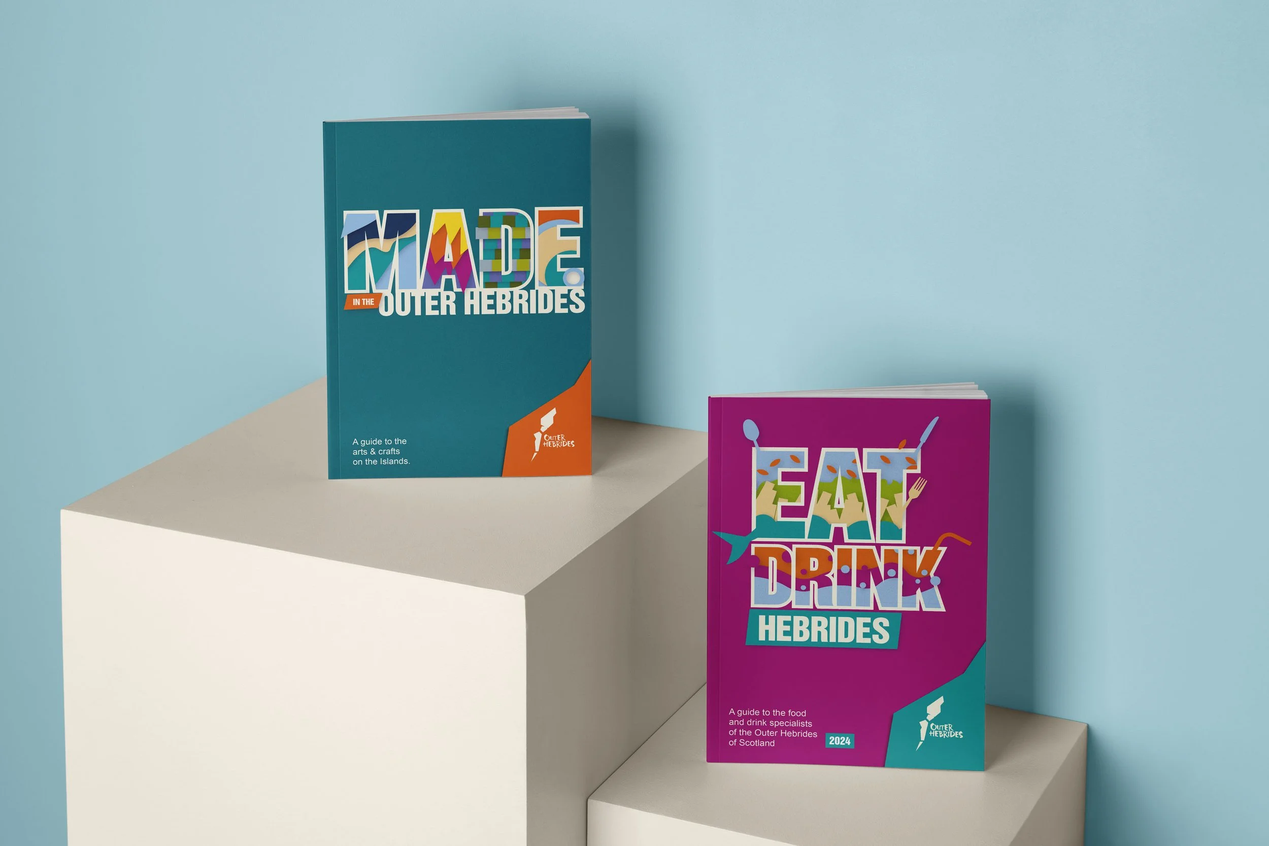 Two colorful guidebooks about the Outer Hebrides displayed on white rectangular blocks, one titled 'Made in the Outer Hebrides' and the other 'Eat, Drink, Hebrides', against a pastel blue background.