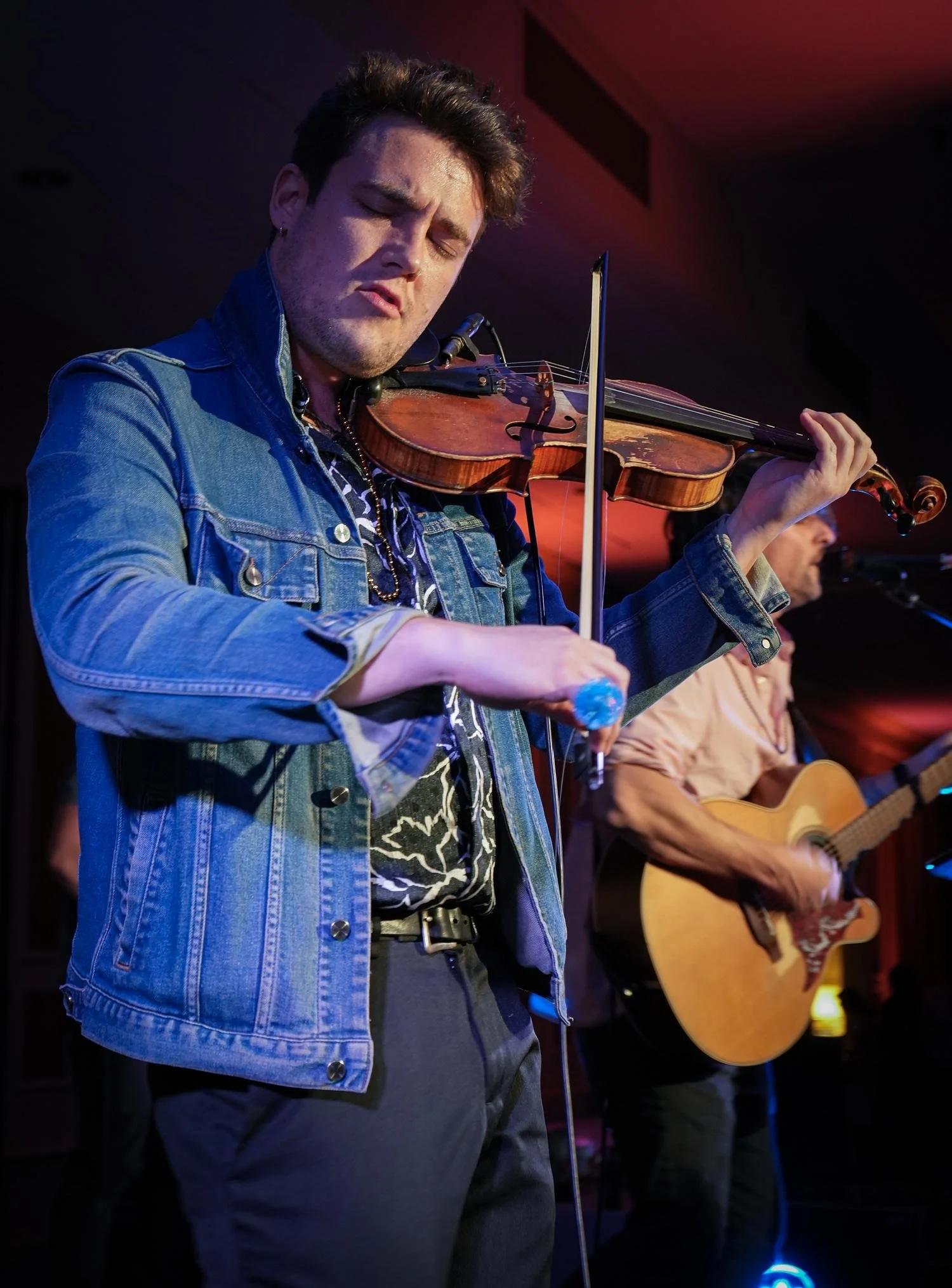 A man with dark hair and a denim jacket playing a violin with his eyes closed, in a dimly lit room with purple and red lighting.