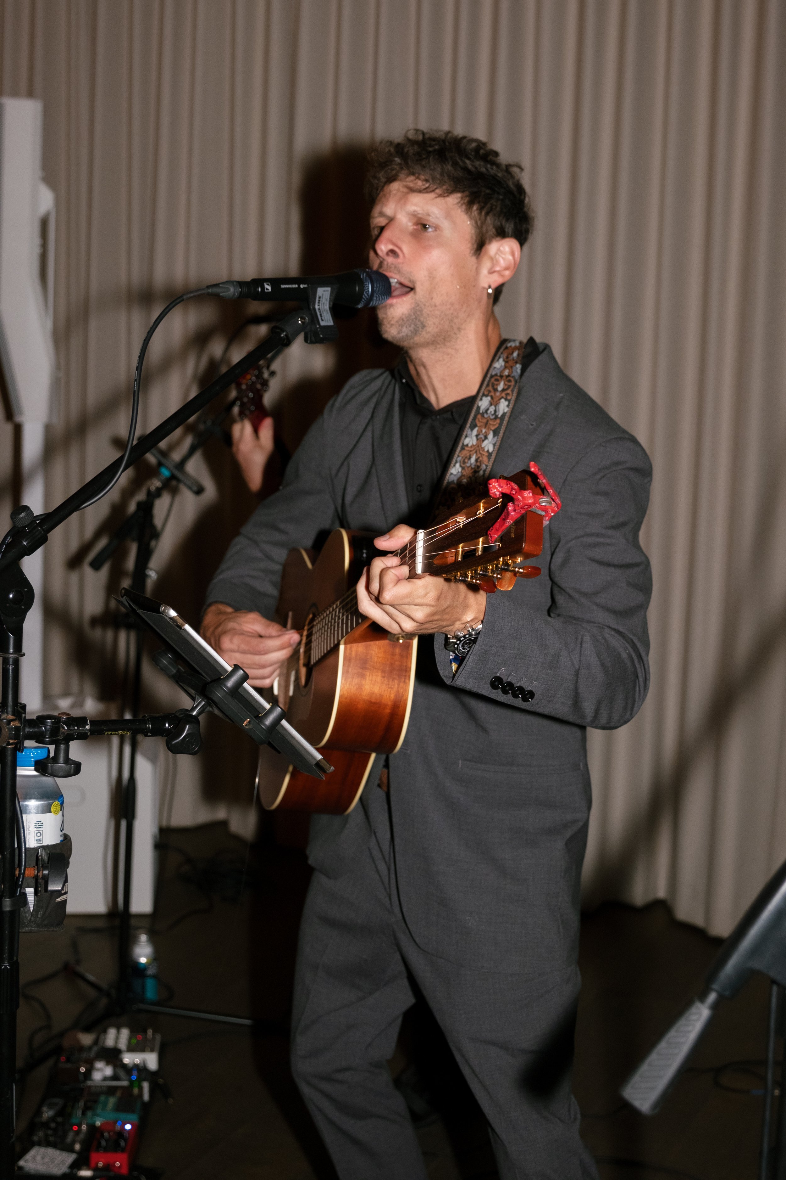 A man in a gray suit playing an acoustic guitar and singing into a microphone during a performance in an indoor setting with beige curtains in the background.