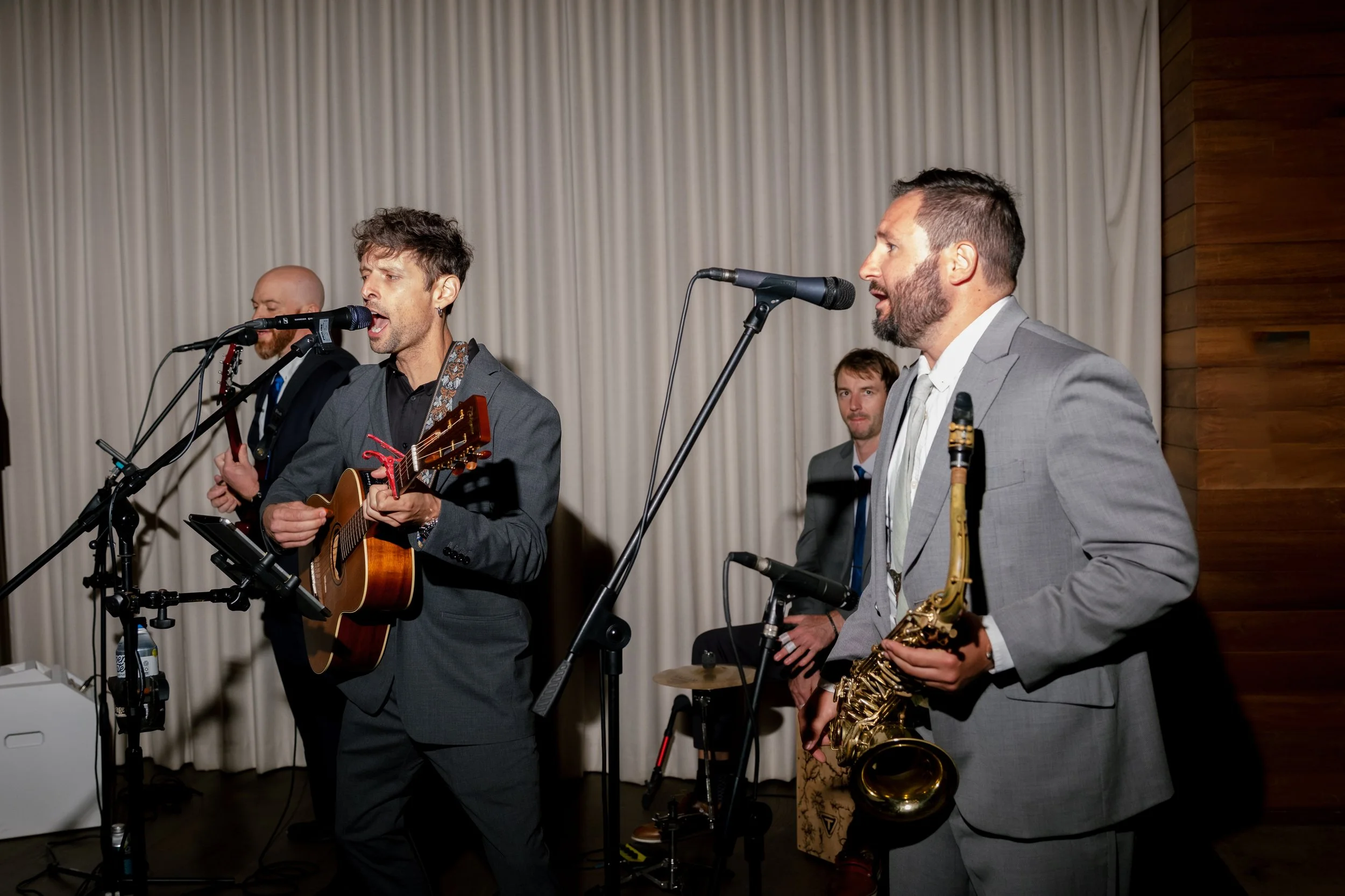 Group of four male musicians performing live. One playing guitar and singing, one holding saxophone, and two in the background, one possibly playing percussion. They are dressed in suits and are set against a backdrop with white curtains and wooden paneling.