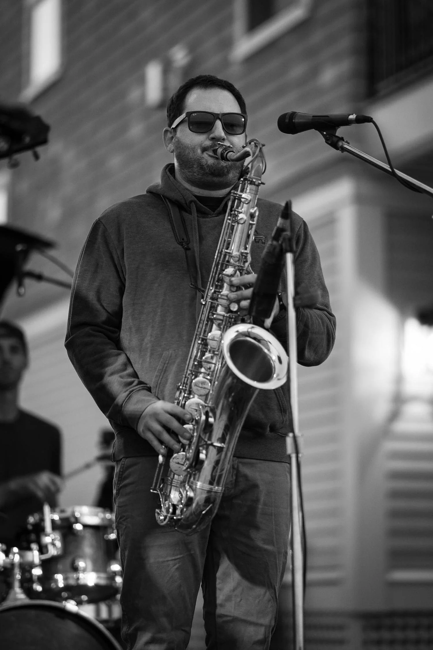 A man wearing sunglasses is playing a saxophone at a music performance, with a microphone in front of him and a drum set visible in the background.