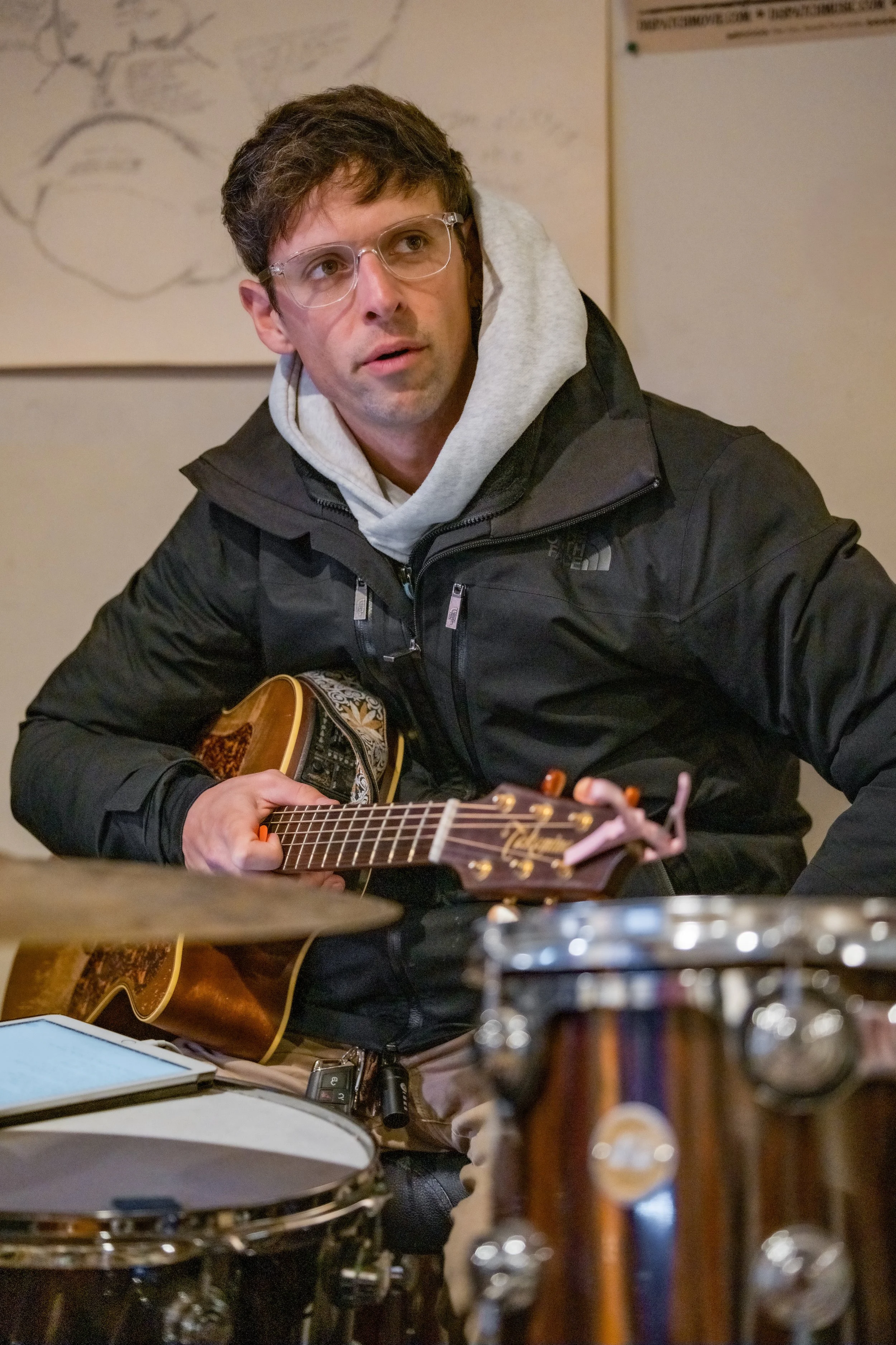 A man with glasses and a hoodie under a black jacket is playing an acoustic guitar. The room has musical instruments including drums in the foreground and background, and a wall with a drawing and a notice.