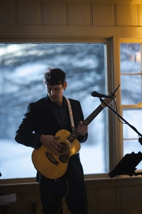 Young man playing an acoustic guitar in front of a window with a snowy landscape outside, during a live performance indoors.