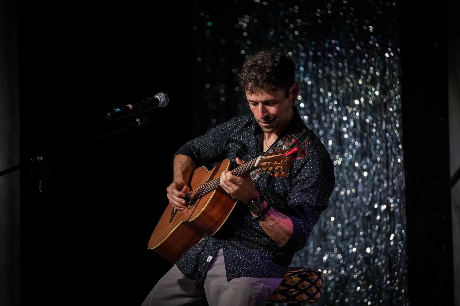 Man playing acoustic guitar on stage with a microphone, dark background with glittering curtain