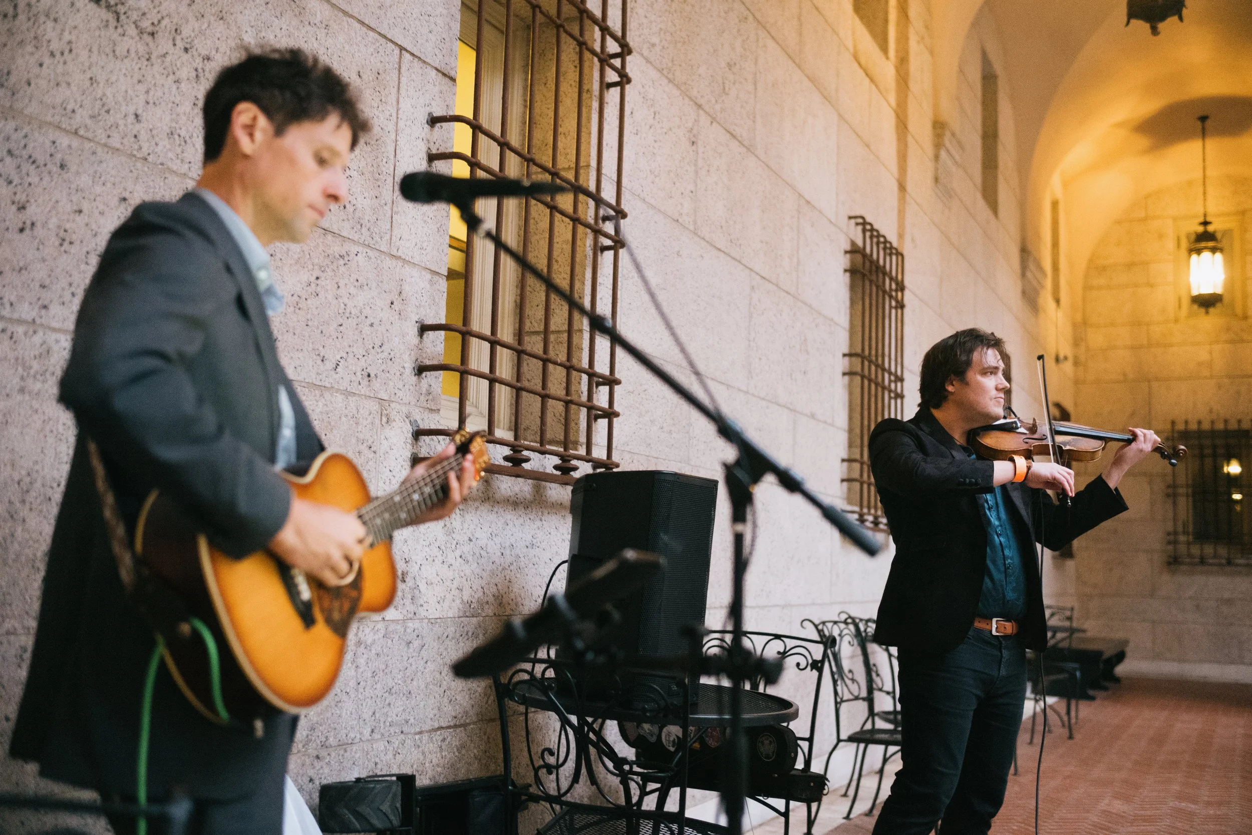 A musician plays guitar while a vocalist sings in an indoor setting with arched ceilings and wall-mounted lamps.