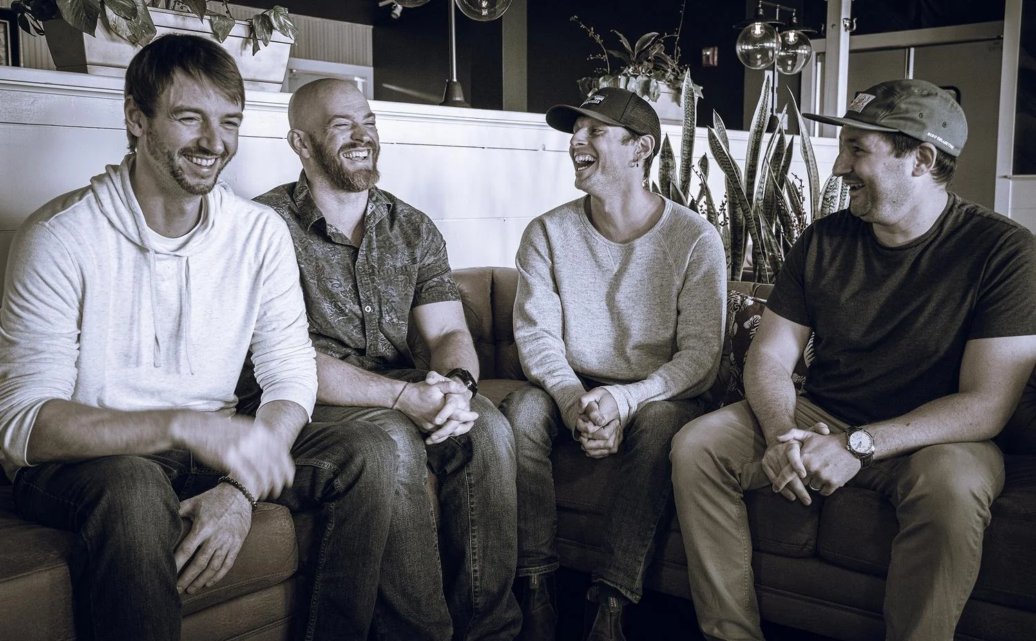 Four friends sitting on a couch, laughing and enjoying conversation in a cozy indoor setting with plants and warm lighting.