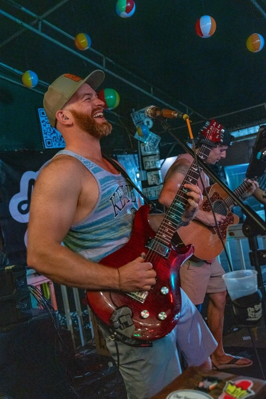 A man with a beard and wearing a tank top and a baseball cap is playing an electric guitar and smiling, while another person in a tank top is playing an acoustic guitar. They are performing at a lively indoor venue decorated with colorful beach balls hanging from the ceiling.
