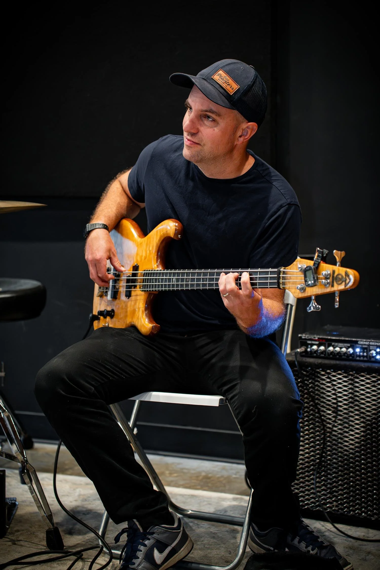 A man sitting and playing an electric guitar in a music rehearsal space. He's wearing a black T-shirt, black pants, athletic shoes, and a backwards black hat with a brown Leather patch. There is a small amplifier nearby, and black soundproof walls in the background.