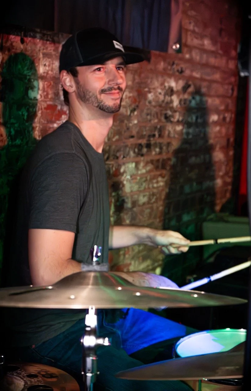 A man wearing a black cap and dark t-shirt playing the drums in a dimly lit venue with a brick wall background.