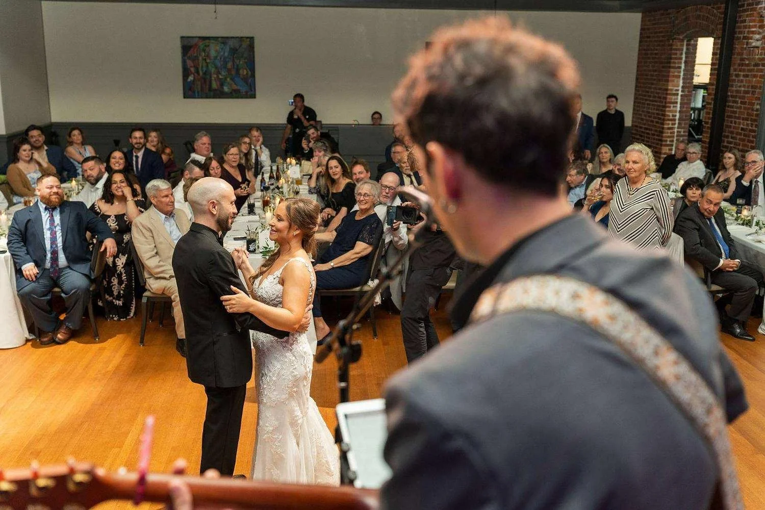 A bride and groom dancing during a wedding reception, surrounded by seated guests who are watching and smiling, with a person in the foreground taking a photo.