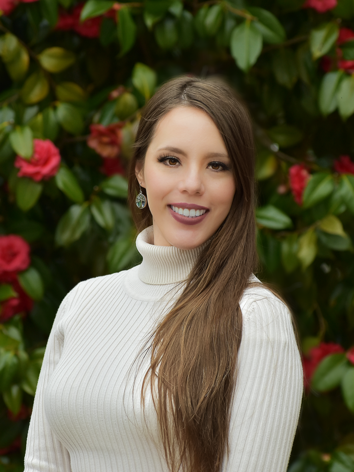 Amanda N. Williams  with long brown hair wearing a white turtleneck sweater and earrings, standing outdoors in front of a bush with pink flowers and green leaves.