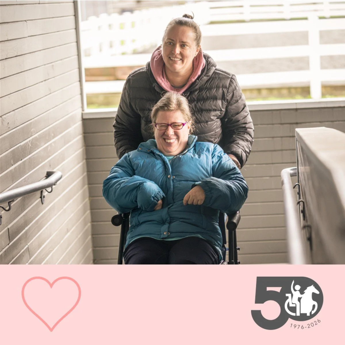 Participant smiles while being pushed in a wheelchair by a staff member along a covered walkway.