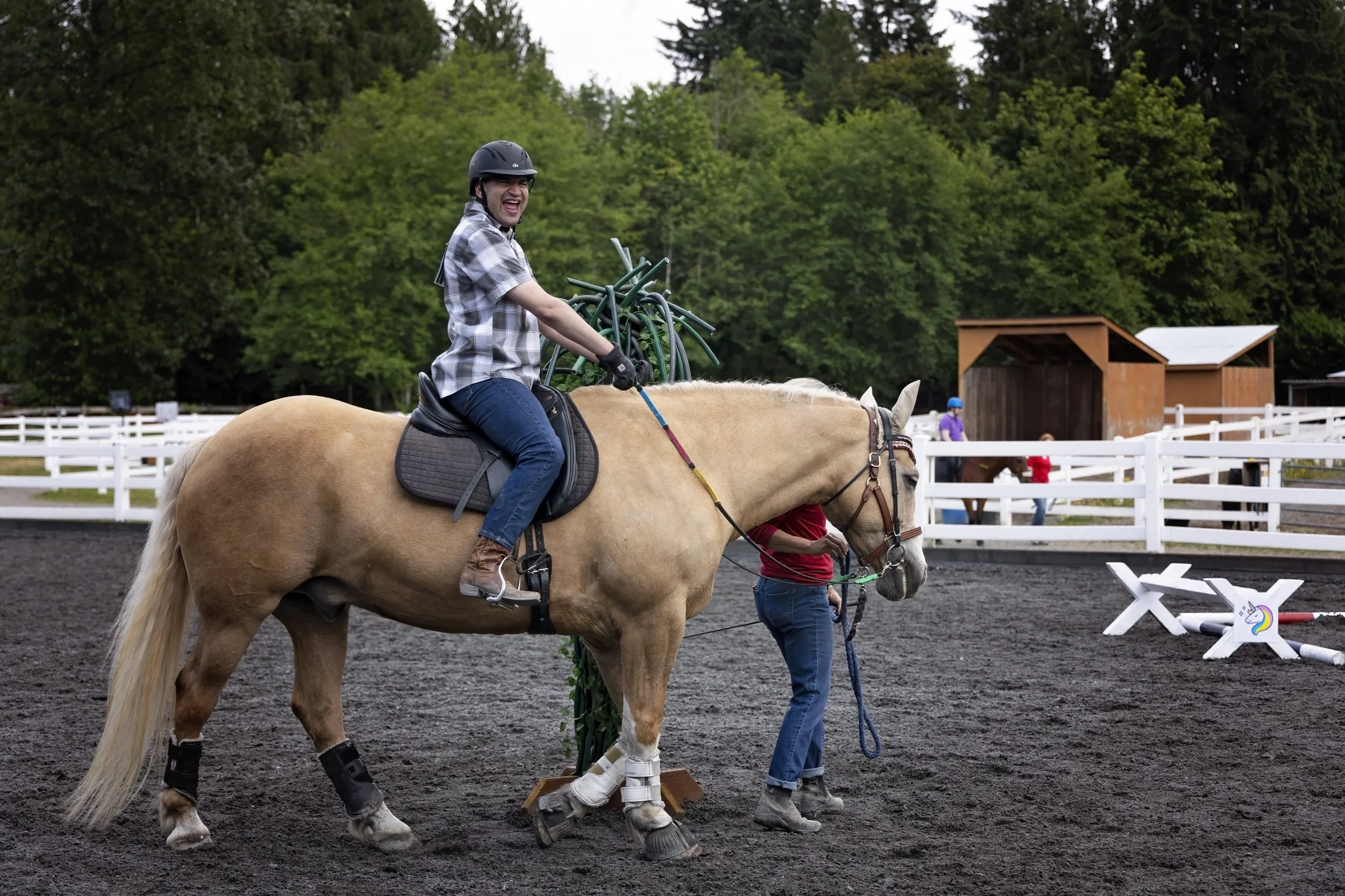 An adaptive rider wearing a helmet smiles while riding a light-colored horse in an outdoor arena. A volunteer walks beside the horse holding the lead, with white fencing, trees, and training obstacles visible in the background.