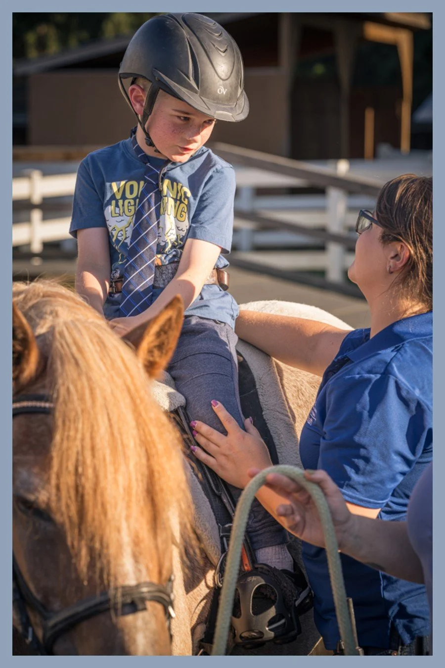 A boy sits on a horse wearing a helmet while a staff member in a blue shirt offers reassurance and adjusts his position.