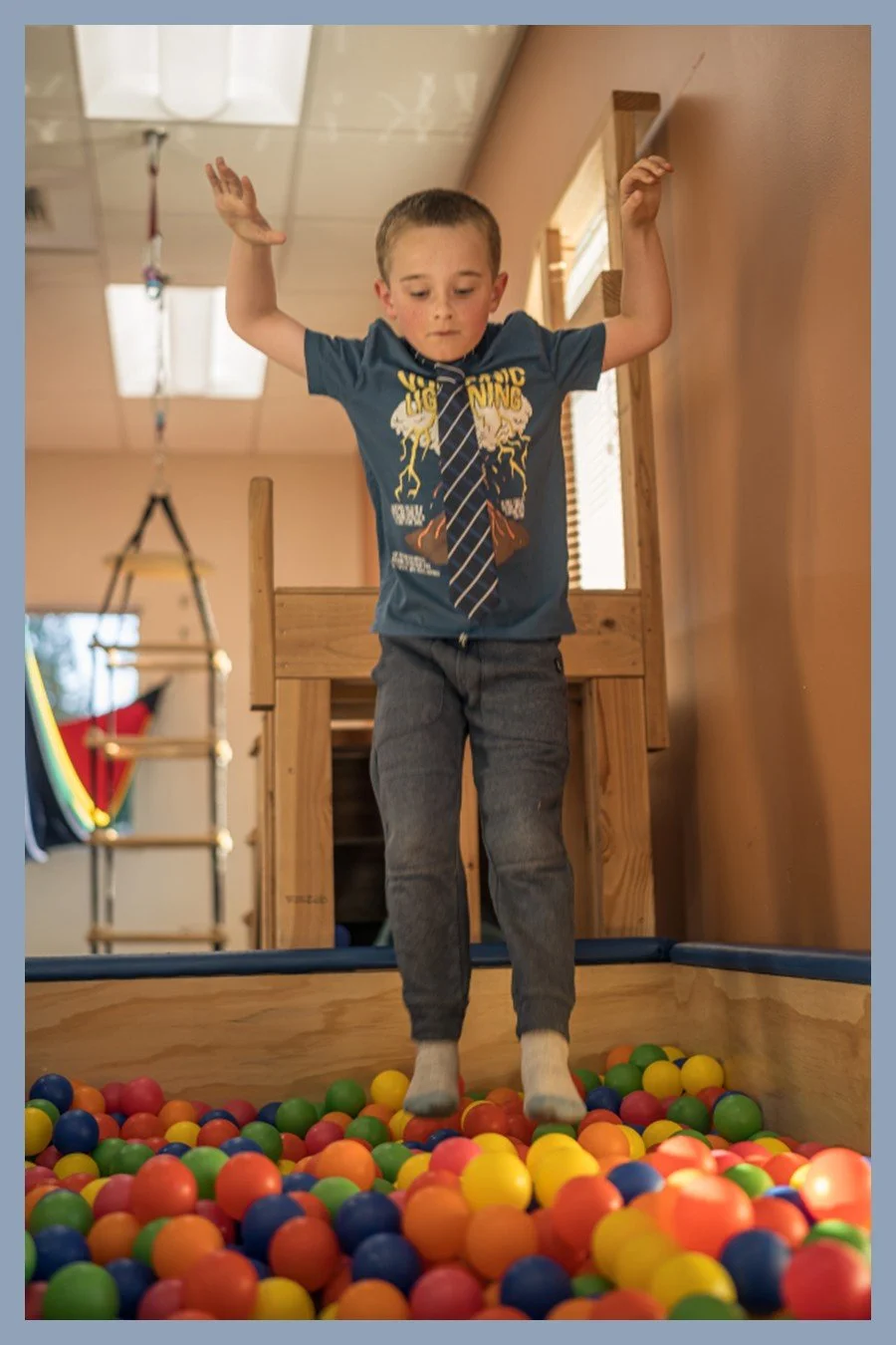 A boy in a blue t-shirt and tie stands poised to jump into a colorful ball pit inside a therapy gym, arms raised for balance.