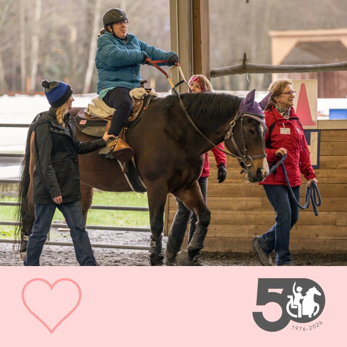 Participant rides a horse indoors while volunteers walk alongside for safety.