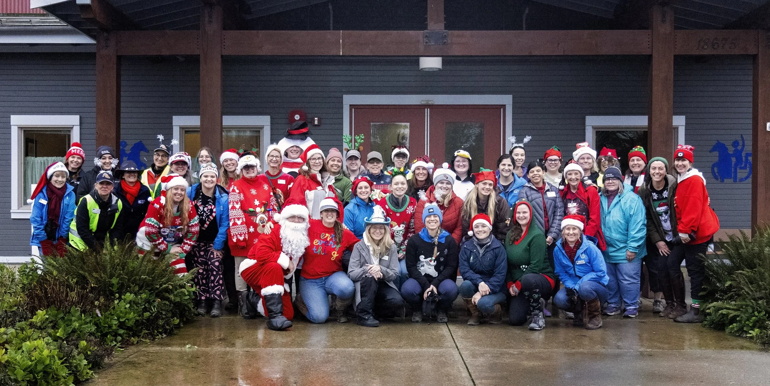 Group of Little Bit staff and volunteers wearing festive holiday hats and sweaters, posing together outside the Welcome Center.