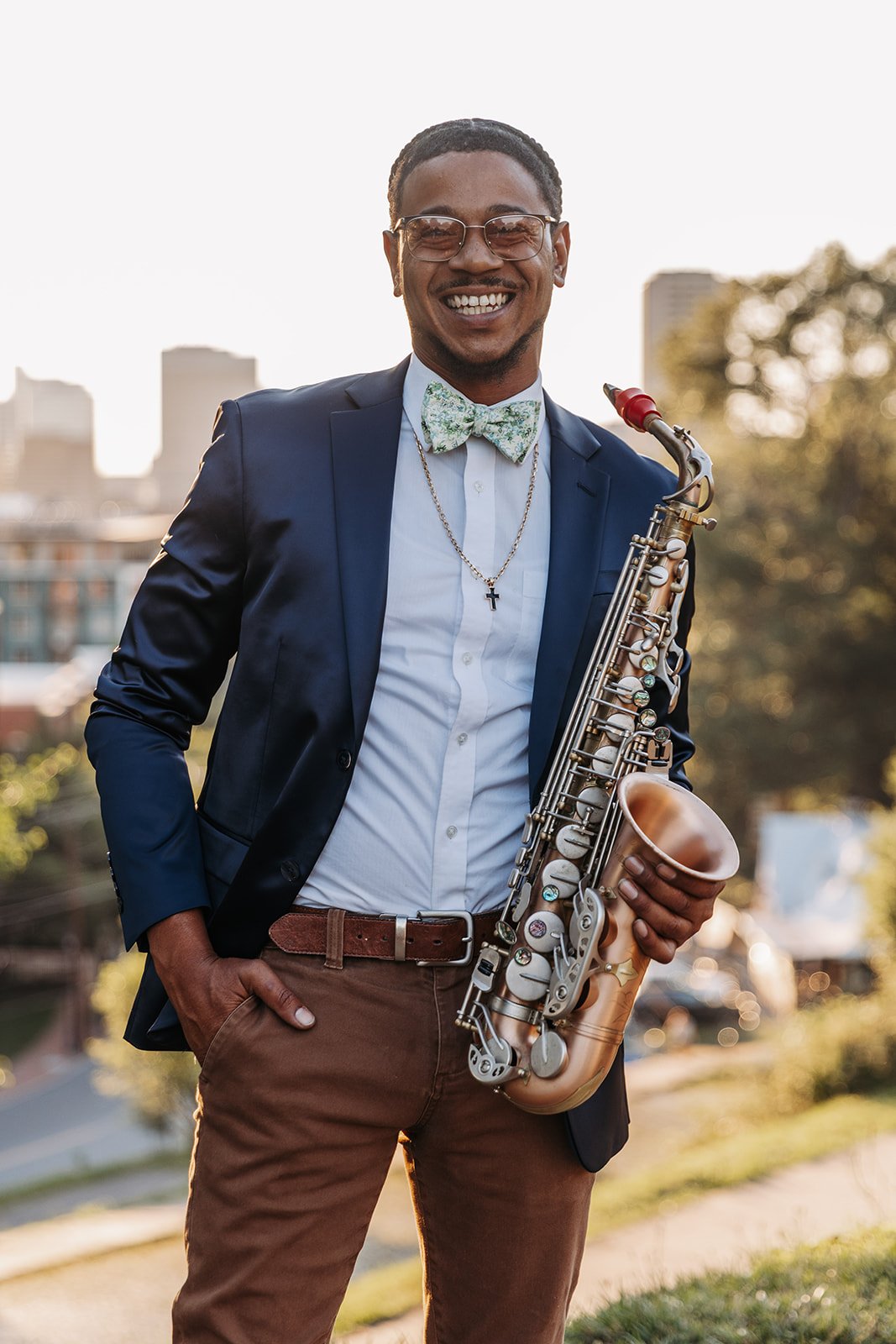 A smiling man in a suit jacket and bow tie holding a saxophone outdoors with city buildings and trees in the background.