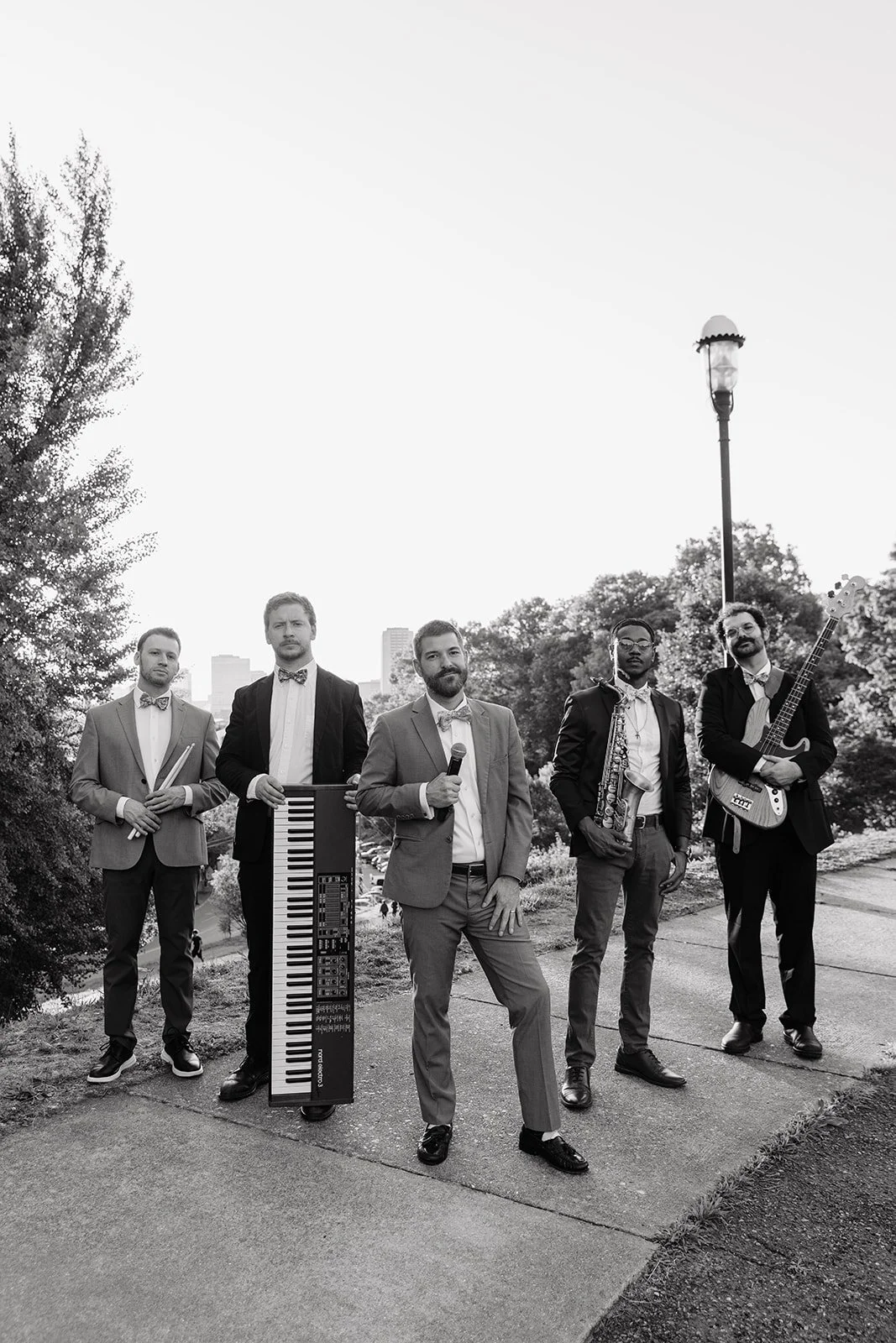 A black and white photo of five men standing outdoors in suits with musical instruments, including a keyboard, saxophone, and guitar, with trees and city buildings in the background.