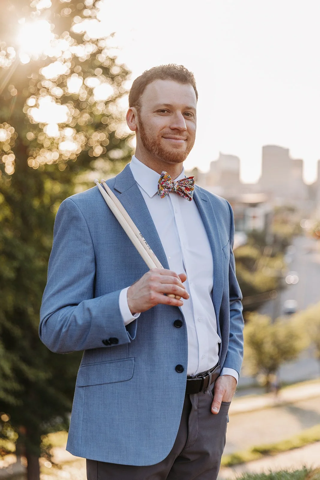 A man in a blue suit with a colorful bow tie, holding a pair of drumsticks, standing outdoors during sunset with buildings in the background.