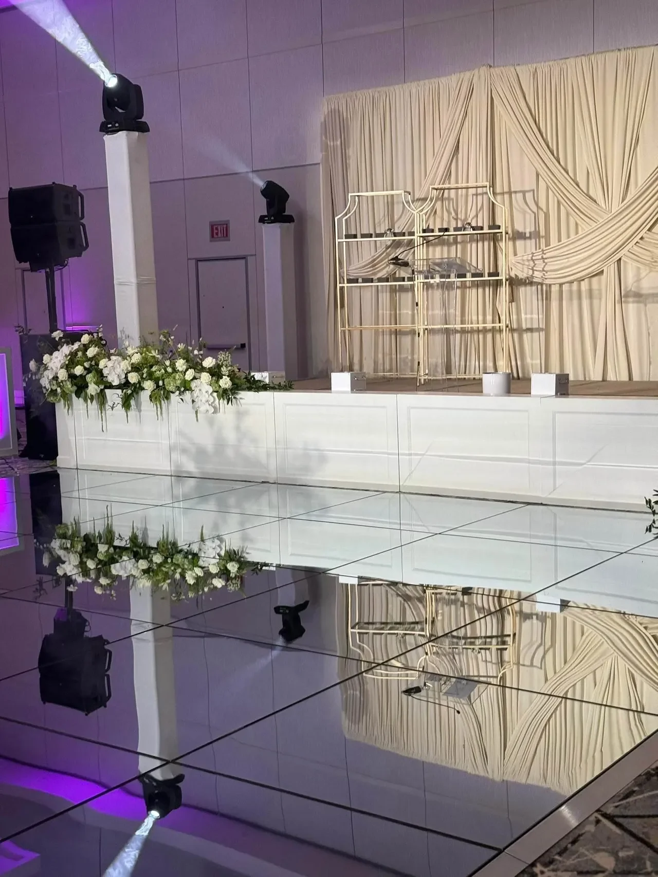 A decorated stage with cream-colored drapes, a floral arrangement in front, and stage lighting, set for a wedding or event.