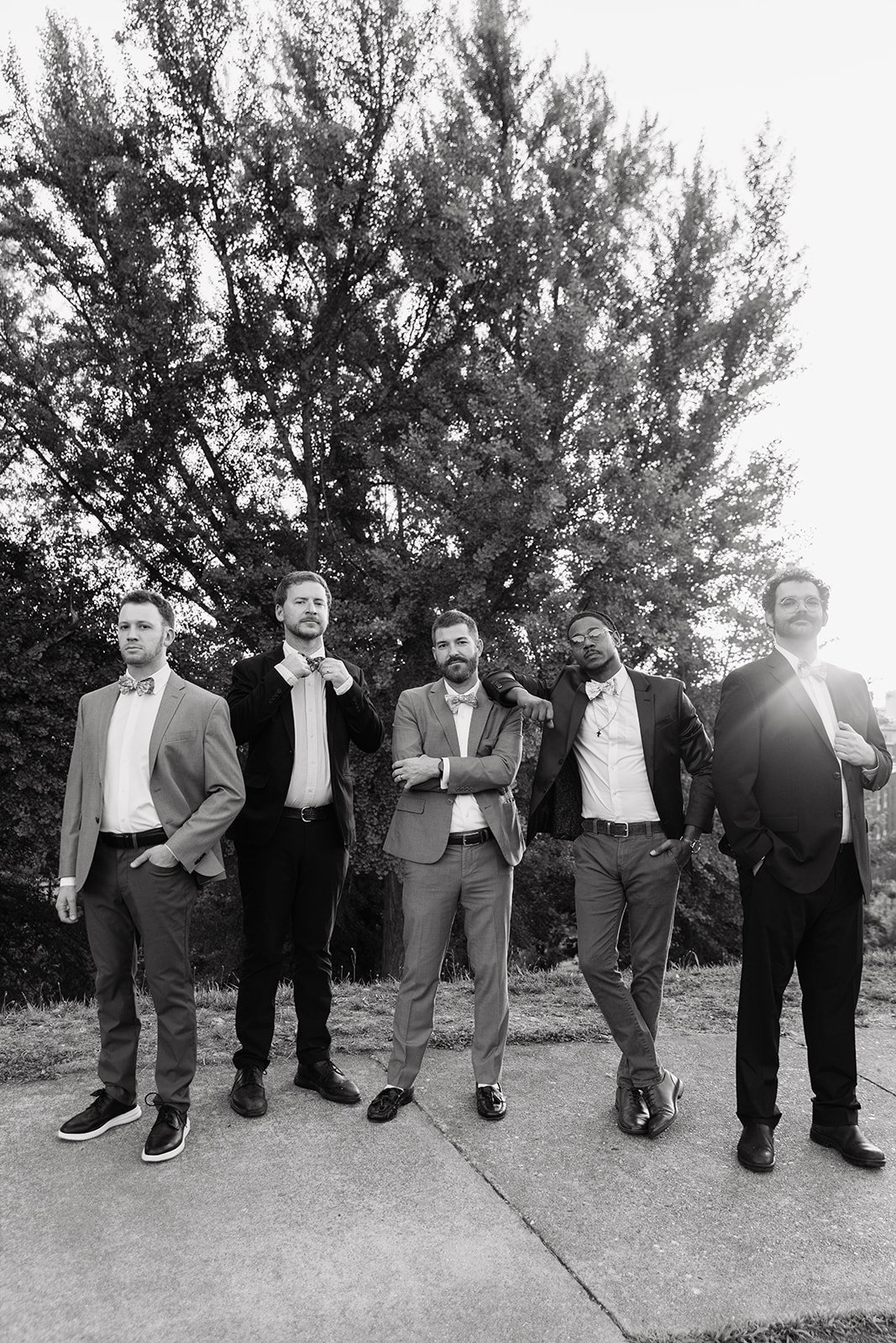 A group of five men dressed in suits and bow ties standing outdoors on a sidewalk in front of a large tree, in black and white, posing for a photo.
