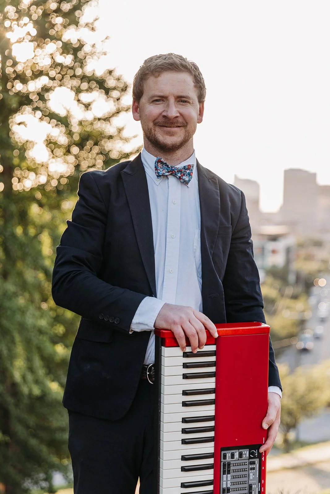 A man in a suit and bow tie holding a red and white keyboard outdoors during sunset.