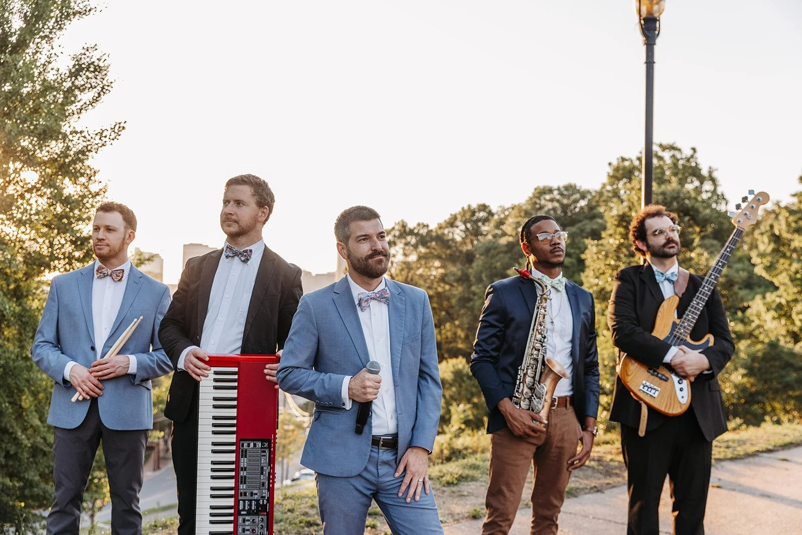 The Bow Tied Band poses outdoors at sunset, featuring a DJ and MC alongside live musicians including drums, keyboard, saxophone, and bass guitar, blending the energy of a live band with the versatility of a wedding DJ for luxury celebrations.