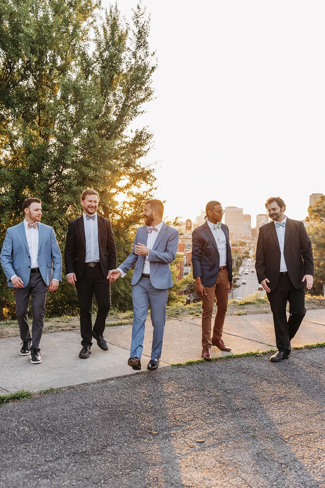 Group of five men dressed in suits, walking and talking outdoors during sunset, with cityscape in the background.