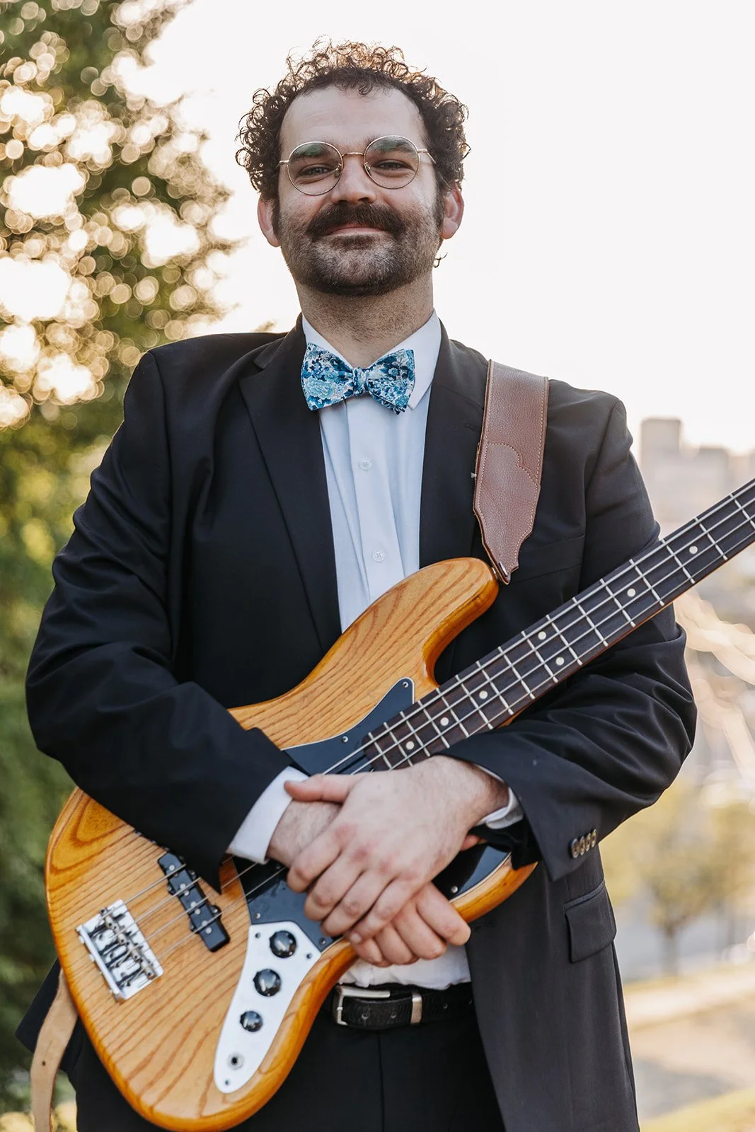 Man in tuxedo with glasses and a blue patterned bow tie holding an electric bass guitar outdoors during sunset.