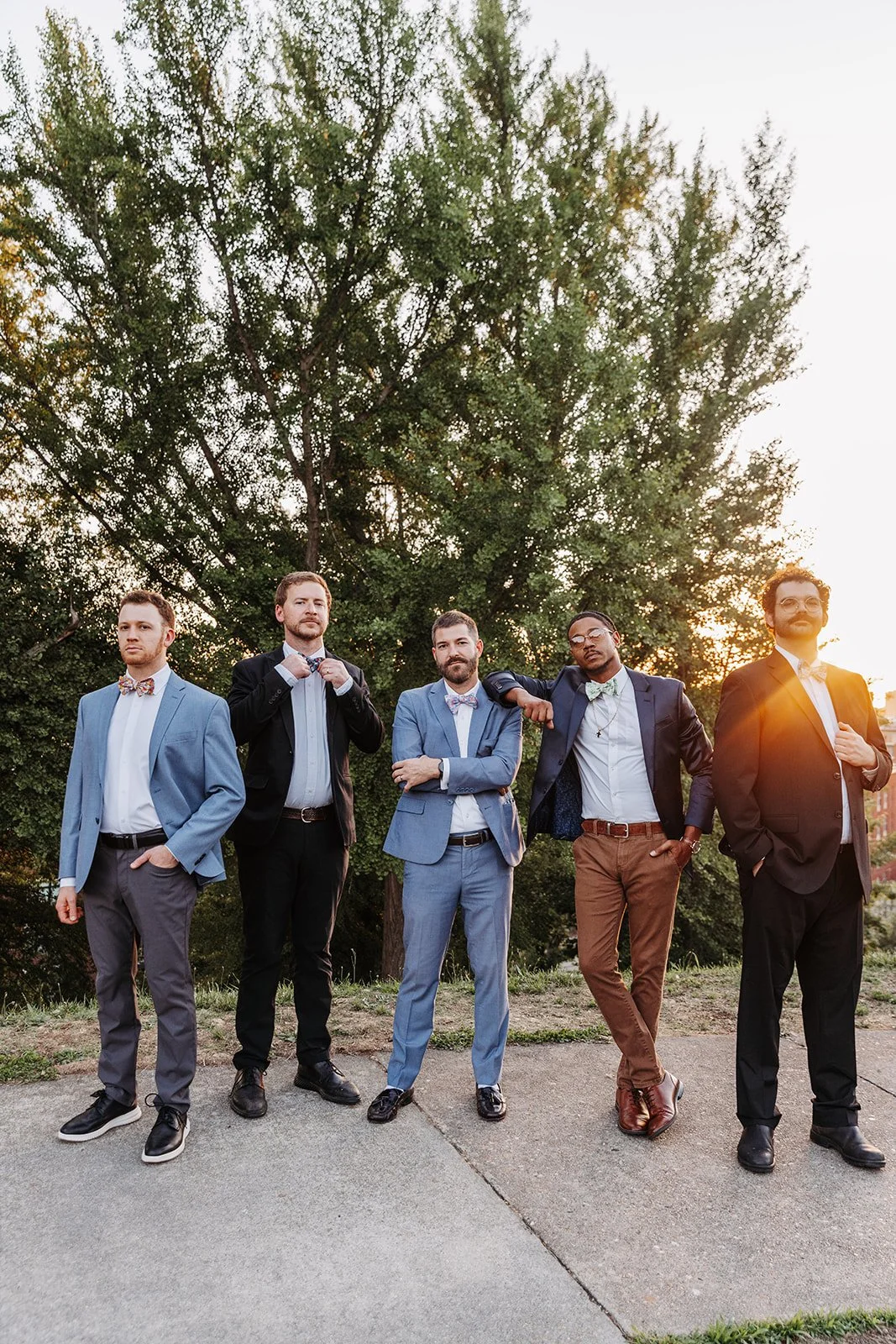 Five men dressed in suits standing outdoors at sunset, with greenery and a large tree behind them.