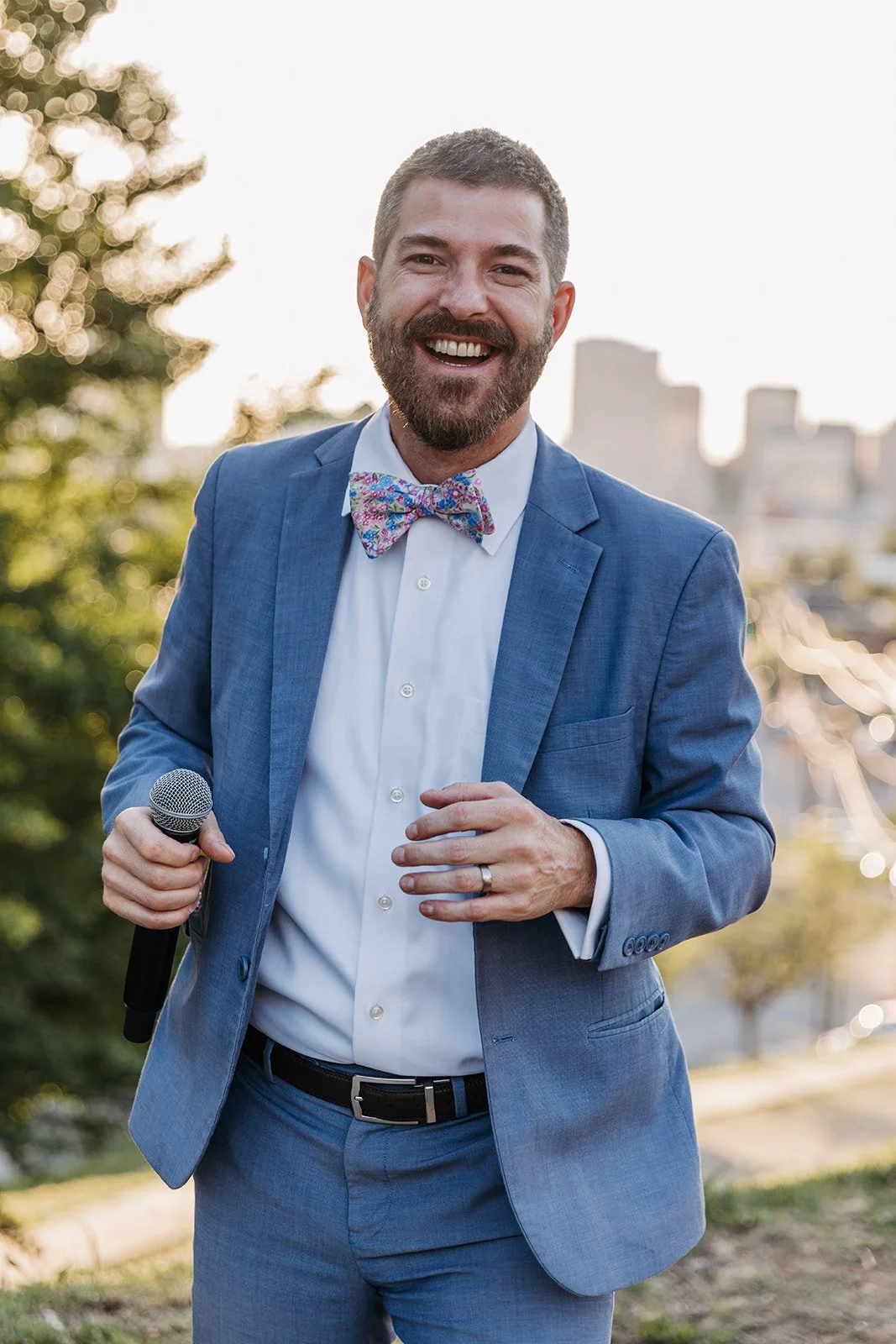A man in a blue suit with a floral bowtie smiling and holding a microphone outdoors with city buildings and trees in the background.