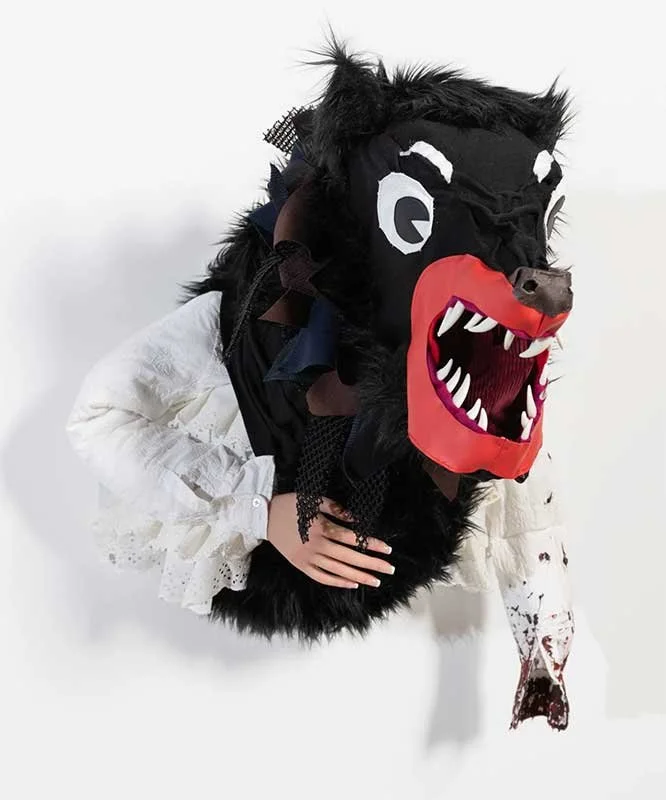 Person wearing a large, black, furry animal mask with big teeth and red lips, dressed in a white, frilly shirt, and emerging from a white background.