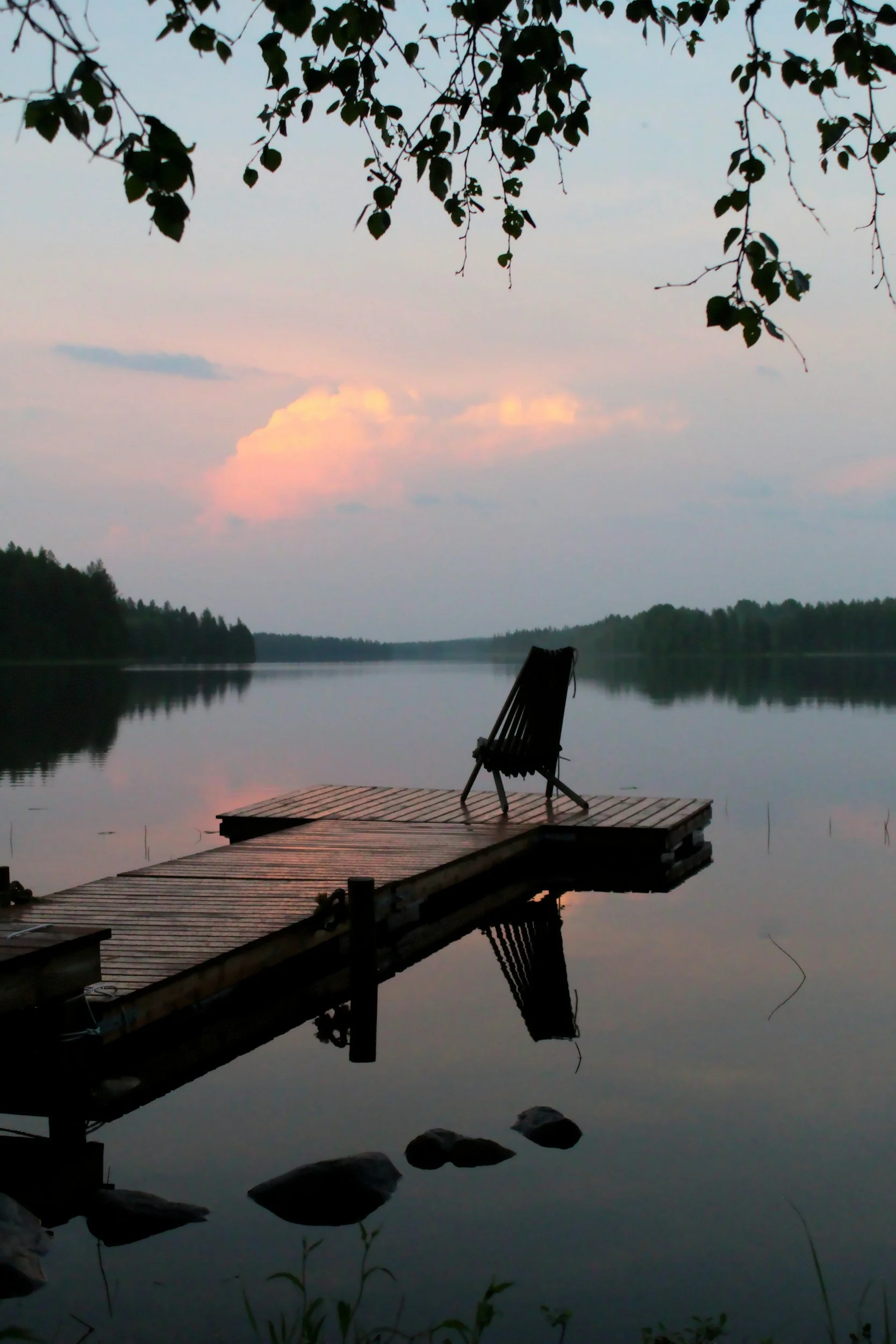 A lake scene at dusk with a wooden dock and a single chair facing the water. The sky has pinkish clouds and is reflected in the calm water, surrounded by trees.