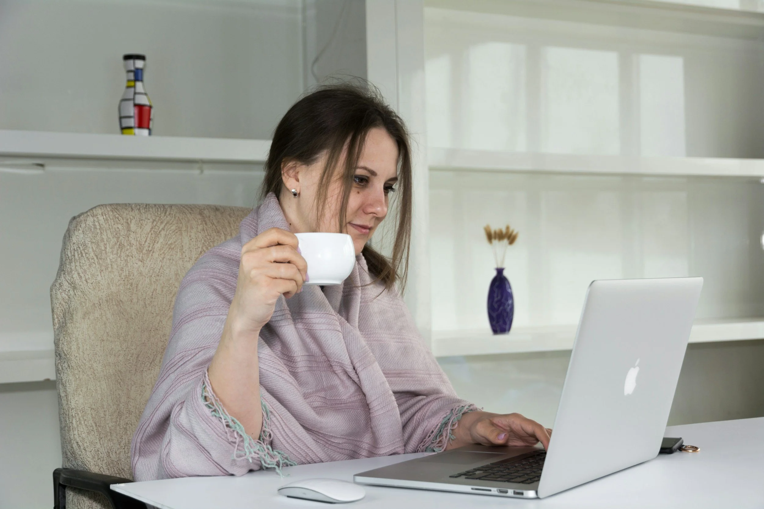 A woman with brown hair sitting at a white desk, holding a white mug, working on a silver laptop, in a bright room with white shelves and a vase with dried flowers.