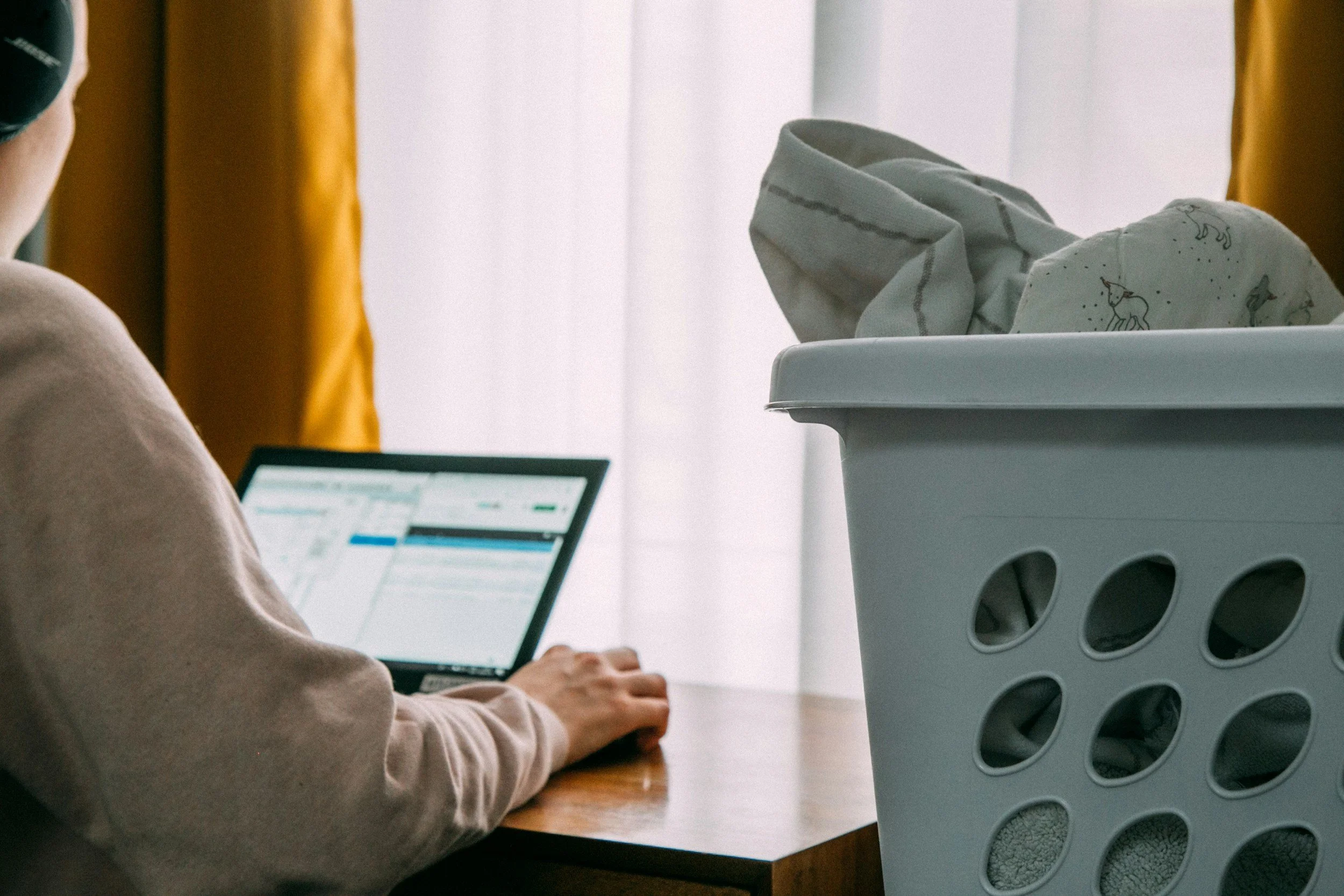 A person working on a laptop with a laundry basket nearby. The background features a window with white curtains and yellow drapes.
