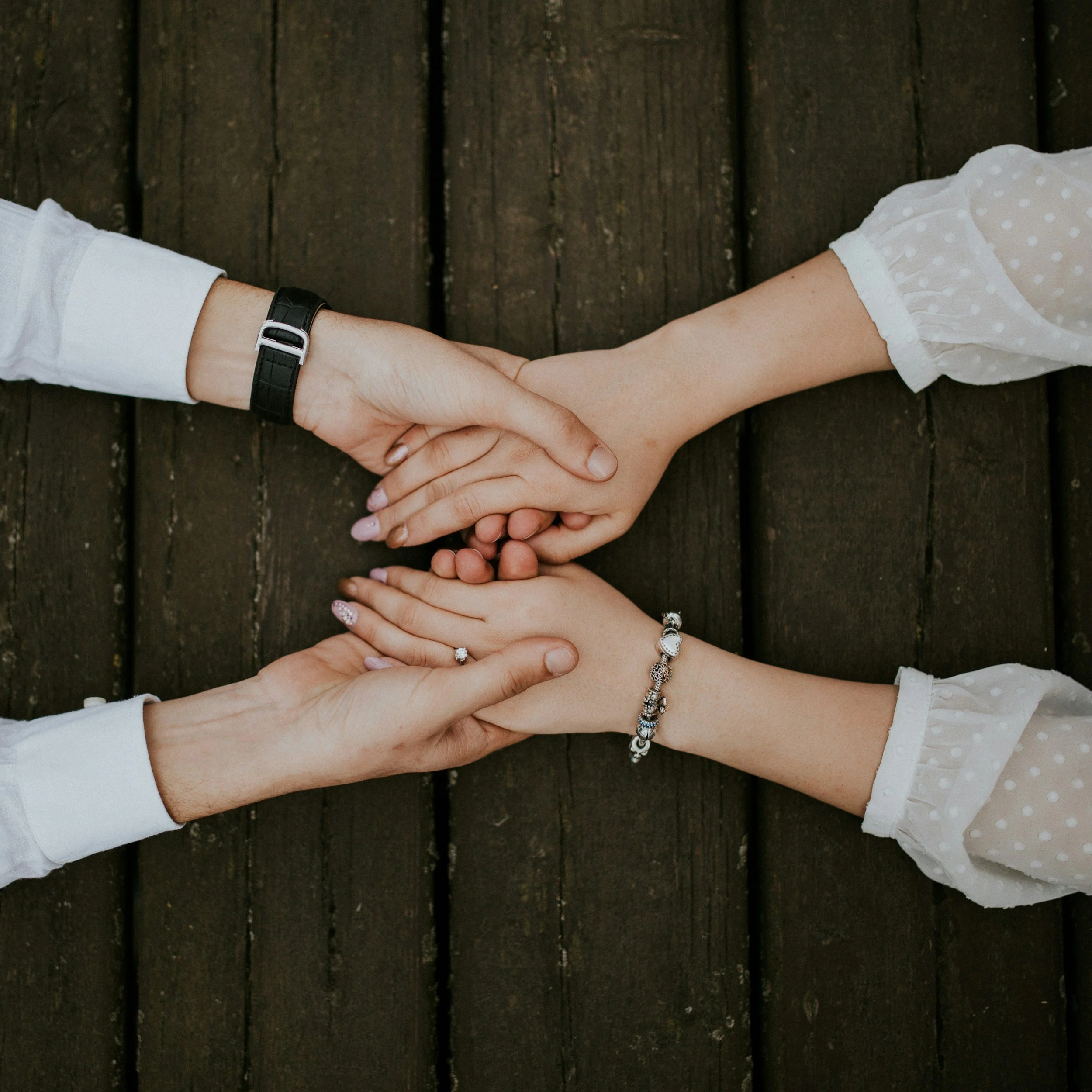 Two people with light skin holding hands on a wooden surface, wearing bracelets and rings.