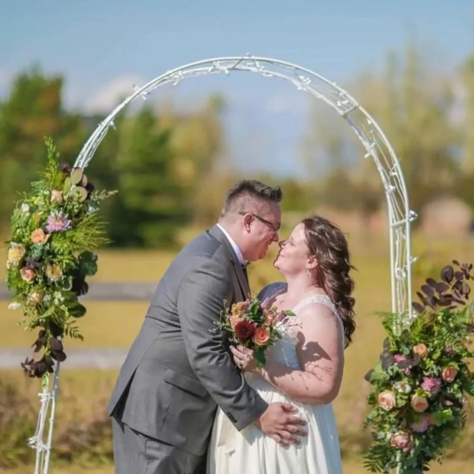 A couple at their wedding, standing under a metal arch decorated with flowers and greenery, touching noses, outdoors with a blurred background of trees and sky.