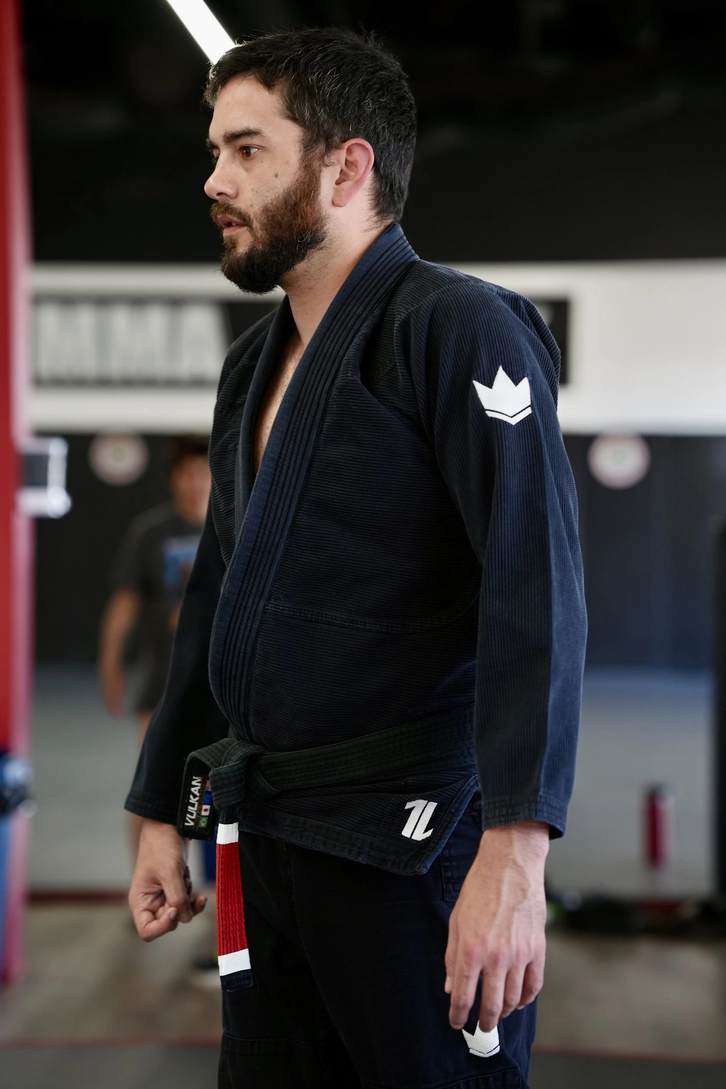 A man with a beard standing in a martial arts gym, wearing a black rash guard with yellow accents and the word 'KINGS' along with other text and designs on the front, in front of a black mat and flags hanging in the background.