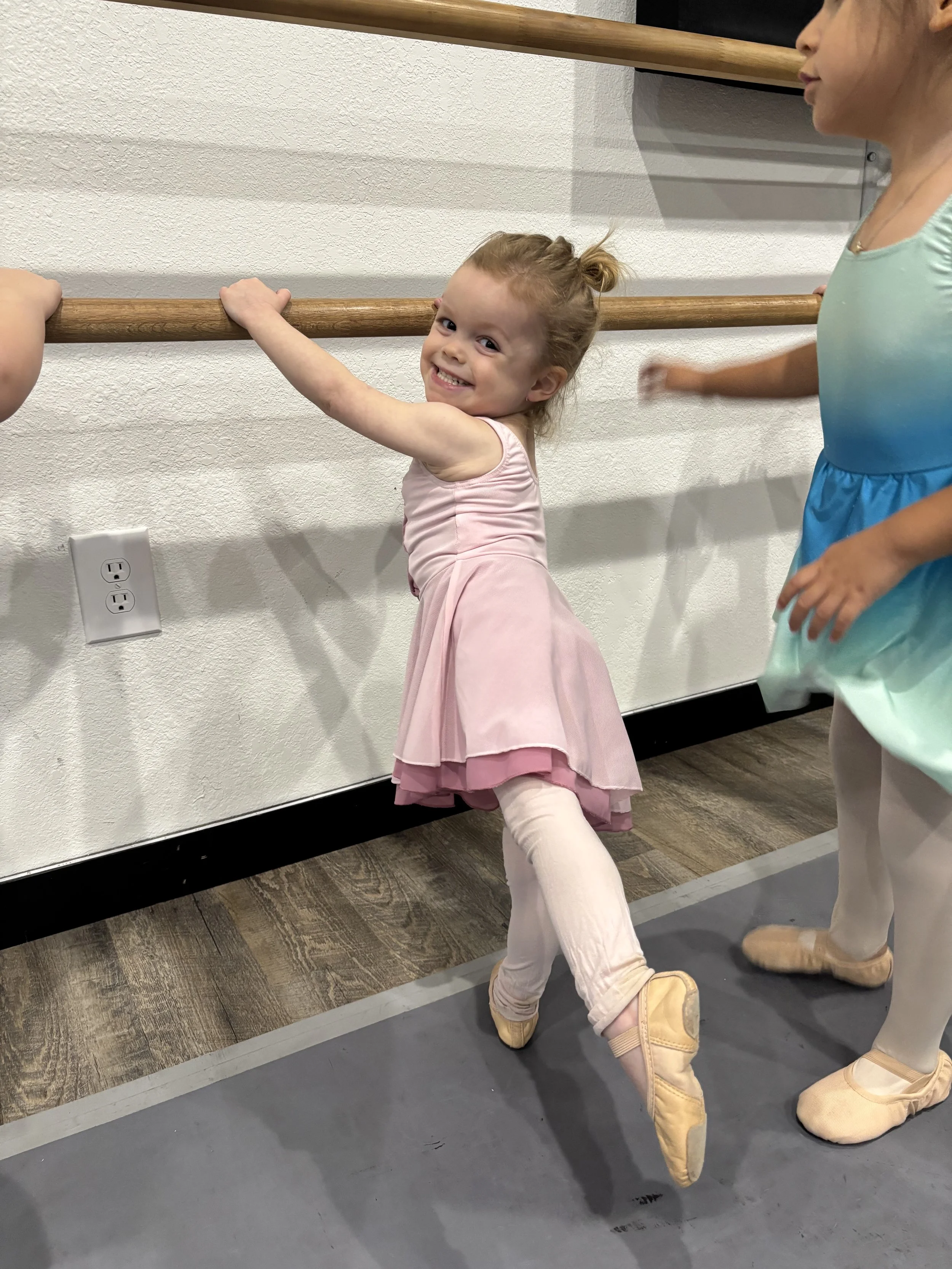 A young girl in a pink ballet dress and tights is smiling while holding onto a ballet barre in a ballet class at miss twister dance company