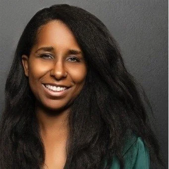 A woman with long, dark hair smiling at the camera against a neutral gray background.