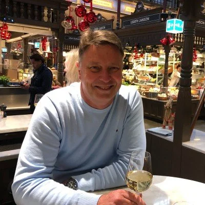 Man sitting at a table in a shopping mall restaurant, smiling, with a glass of white wine, decorated with holiday ornaments.