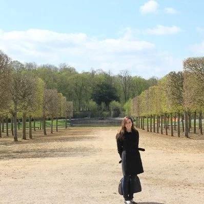 A woman standing on a wide pathway in a park with lined trees and green grass, under a partly cloudy sky.