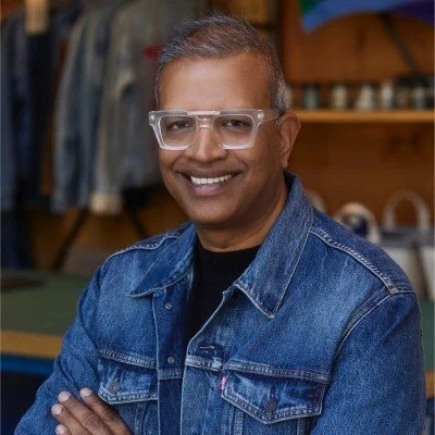 Smiling man wearing safety glasses and a denim jacket in a workshop setting.