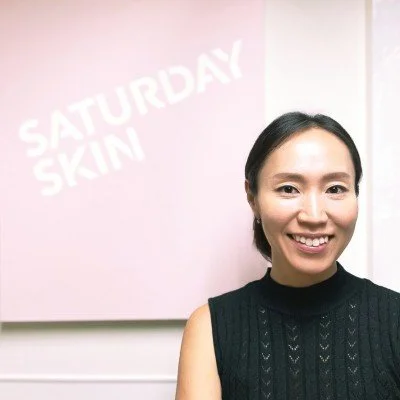 A woman with black hair smiling in front of a pink wall with white text that reads 'SATURDAY SKIN'.