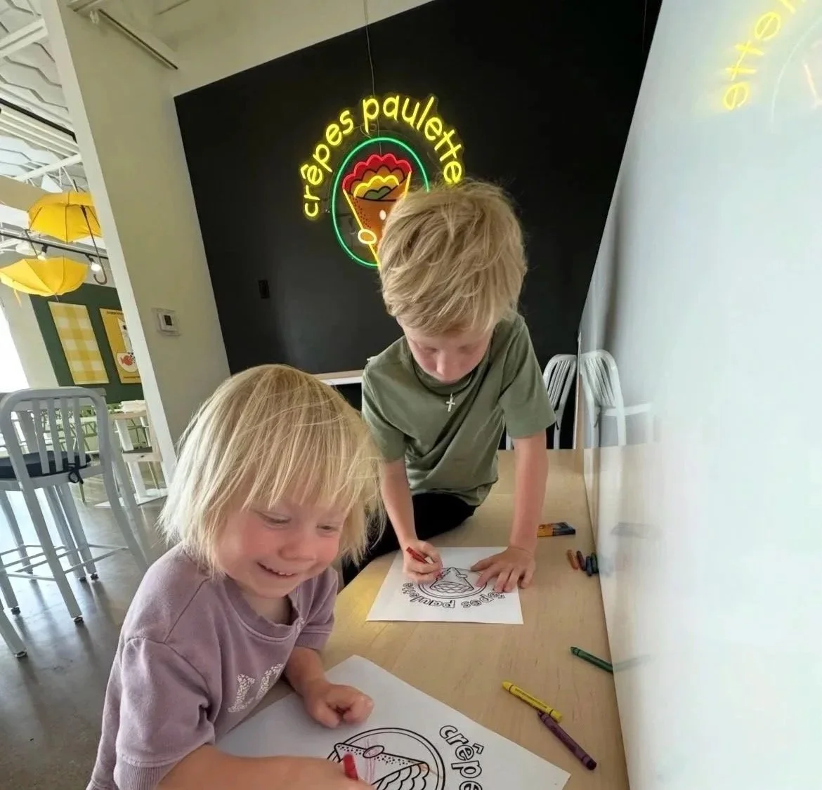 two small children shown coloring on a bench in front of a neon Crepes Paulette sign