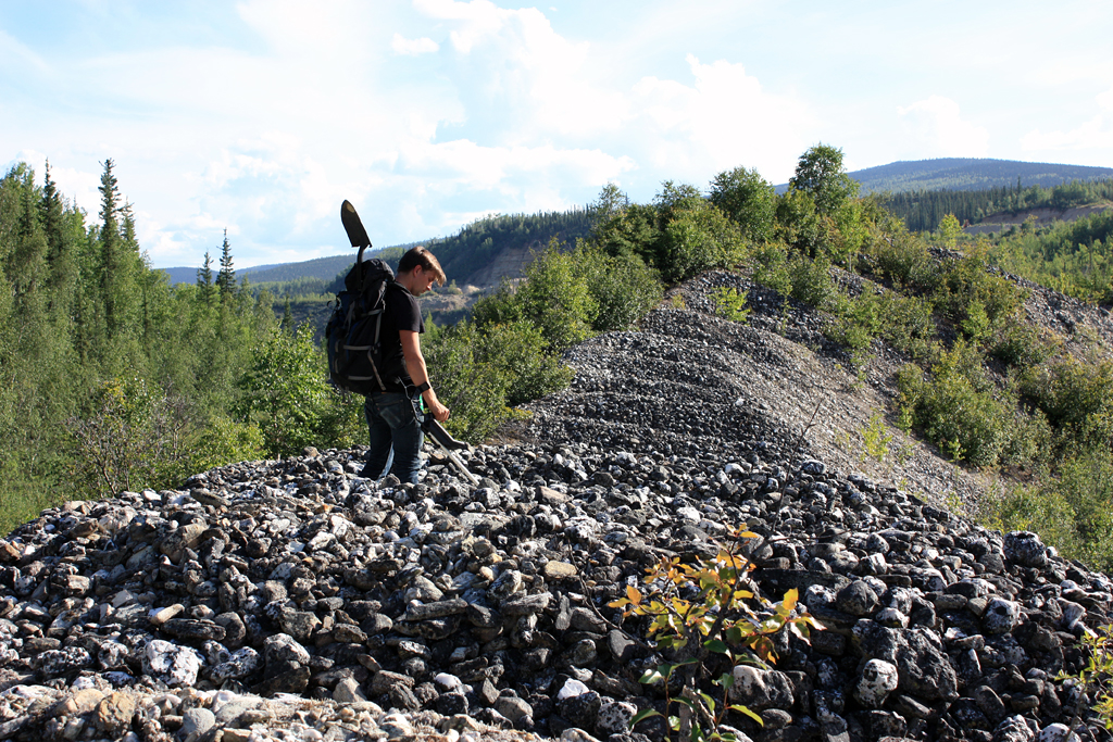 Time Machine for Abandoned Futures (2015) During my first visit to Dawson City, in January 2010, I was brought to look out at the vast intestinal tailing piles left by decades of dredging in the area. For me, this vista became the lasting image of my trip to the Yukon. In a town that boasts many historical monuments, these tailings seemed to represent the ‘authentic’ ruins of the Klondike gold rush. In revisiting Dawson, I began with a quintessentially Klondike activity – a kind of treasure hunting. Over the course of several weeks, I walked along the dredge tailings near Bonanza Creek, using a metal detector to excavate industrial cast-offs. With this collection in tow, I brought it to the Midnight Dome, where I installed my Time Machine ; a strange off-the-grid laboratory which became my home for over a week. This shelter adopts design strategies common in earthship architecture (rain-water collection, south facing greenhouse windows, rammed earth, etc.), but rather than environmental sustainability as its guiding principle, this bubbling chemical structure comes closer to the absurd inefficiency of many of our modern industrial pursuits. Powering this machine is a massive, roof-top battery, in which etching plates and etching acid power an electrolytic cleaning process to remove the rust from my scavenged artifacts. Once cleaned, I meticulously etched the markings left by decades of rust and erosion, forming a kind of topographical map. The result is a glistening surface that memorializes the artifact’s entire lifespan. Overlooking the dredge tailings, this machine presents a kind of prototype for the preservation of degradation. Time Machine for Abandoned Futures was produced with the support of Klondike Institute of Art & Culture and Canada Council for the Arts Image: collecting artifacts near Bonanza Creek, 2015.