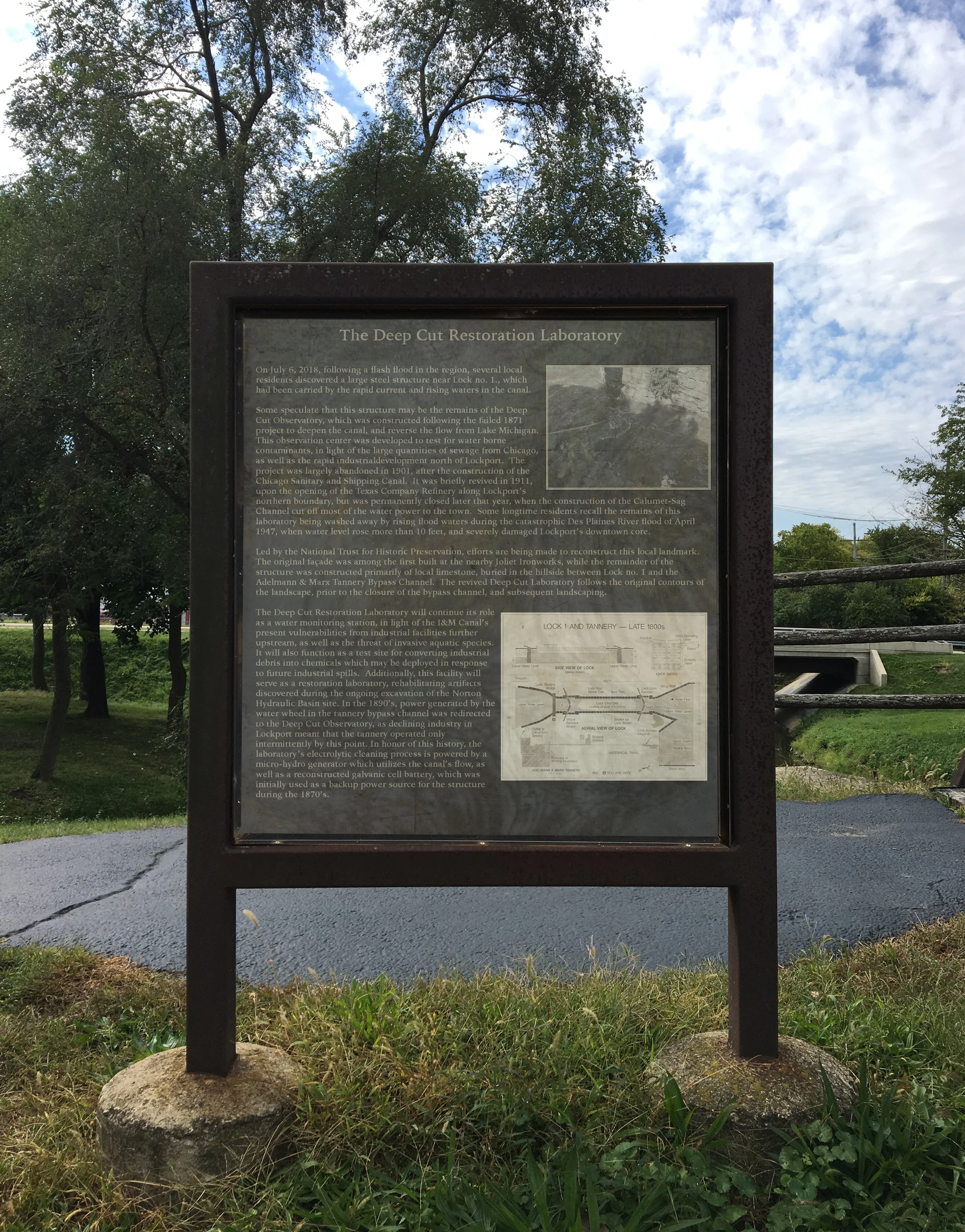 Proposed Historic Signage at I&M Canal Lock no. 1, Lockport, IL Full Text: On July 6, 2018, following a flash flood in the region, several local residents discovered a large steel structure near Lock no. 1., which had been carried by the rapid current and rising waters in the canal. Some speculate that this structure may be the remains of the Deep Cut Observatory, which was constructed following the failed 1871 project to deepen the canal, and reverse the flow from Lake Michigan. This observation center was developed to test for water borne contaminants, in light of the large quantities of sewage from Chicago, as well as the rapid industrial development north of Lockport. The project was largely abandoned in 1901, after the construction of the Chicago Sanitary and Shipping Canal. It was briefly revived in 1911, upon the opening of the Texas Company Refinery along Lockport’s northern boundary, but was permanently closed later that year, when the construction of the Calumet-Sag Channel cut off most of the water power to the town. Some longtime residents recall the remains of this laboratory being washed away by rising flood waters during the catastrophic Des Plaines River flood of April 1947, when water level rose more than 10 feet, and severely damaged Lockport’s downtown core. Led by the National Trust for Historic Preservation, efforts are being made to reconstruct this local landmark. The original façade was among the first built at the nearby Joliet Ironworks, while the remainder of the structure was constructed primarily of local limestone, buried in the hillside between Lock no. 1 and the Adelmann & Marx Tannery Bypass Channel. The revived Deep Cut Laboratory follows the original contours of the landscape, prior to the closure of the bypass channel, and subsequent landscaping. The Deep Cut Restoration Laboratory will continue its role as a water monitoring station, in light of the I&M Canal’s present vulnerabilities from industrial facilities further upstream, as well as the threat of invasive aquatic species. It will also function as a test site for converting industrial debris into chemicals which may be deployed in response to future industrial spills. Additionally, this facility will serve as a restoration laboratory, rehabilitating artifacts discovered during the ongoing excavation of the Norton Hydraulic Basin site. In the 1890’s, power generated by the water wheel in the tannery bypass channel was redirected to the Deep Cut Observatory, as declining industry in Lockport meant that the tannery operated only intermittently by this point. In honor of this history, the laboratory’s electrolytic cleaning process is powered by a micro-hydro generator which utilizes the canal’s flow, as well as a reconstructed galvanic cell battery, which was initially used as a backup power source for the structure during the 1870’s.