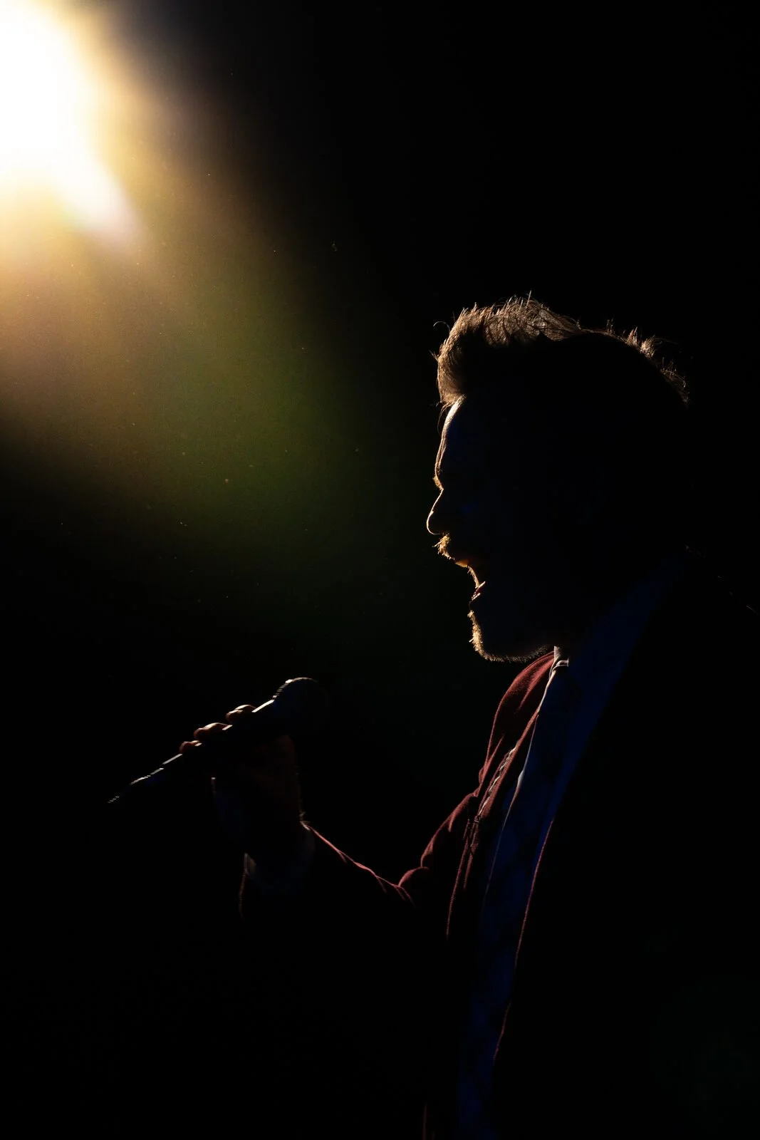 Silhouette of a man singing into a microphone with a bright light shining from the upper left corner.