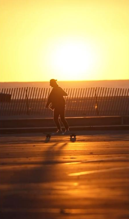 Person skateboarding at sunset on a boardwalk with a fence and ocean in the background.